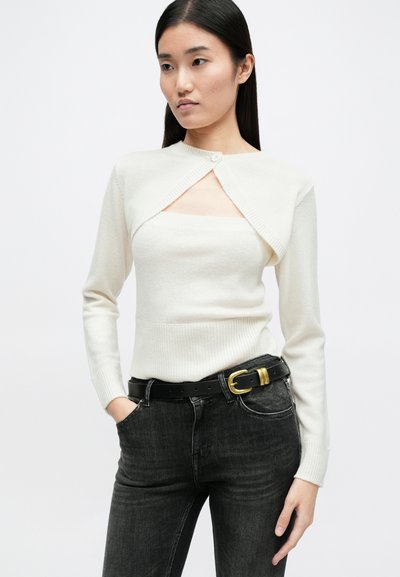 Λευκό πλεκτό πουλόβερ με κλείσιμο με κουμπί, διαθέτοντας σχέδιο με κοψίματα και εφαρμοστά μανίκια, σε συνδυασμό με μαύρα τζιν και μαύρη ζώνη.