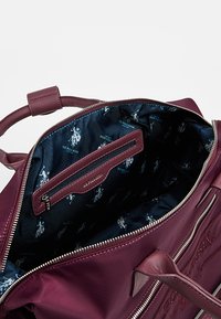 Viininpunainen nahkainen käsilaukku hopeisella vetoketjulla, sisäpuolella tumma kangas, jossa toistuva logokuvio, sisältää vetoketjullisen taskun.