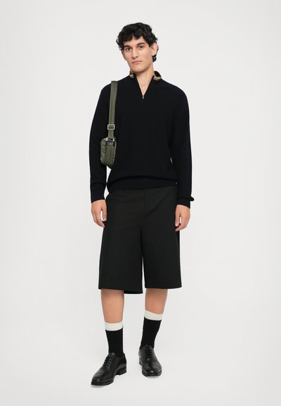 Maglione nero lavorato a maglia con chiusura a zip e un colletto multicolore, abbinato a pantaloni corti neri fino al ginocchio, calze nere e scarpe stringate nere.