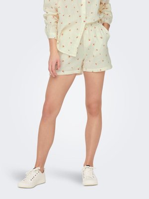 Vrouw draagt crèmekleurig bloemenprint shirt met lange mouwen en bijpassende shorts met witte sneakers, handen in de zakken, staand op witte achtergrond.