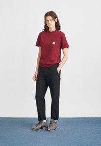 T-shirt bordeaux a maniche corte con tasca sul petto, abbinato a pantaloni cargo neri e scarpe da trekking grigie su pavimento blu.