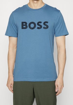 Mężczyzna w niebieskiej koszulce z krótkim rękawem z wyraźnym czarnym napisem "BOSS" na klatce piersiowej i ciemnozielonych spodniach, stojący na jednolitym tle.