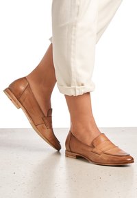 Mocasines de cuero marrón con un tacón bajo, que presentan un diseño elegante y acentos de costura, combinados con pantalones blancos arremangados.