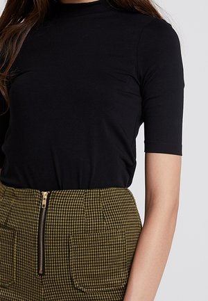 Kobieta ubrana w czarny krótki top wsunięty w oliwkowozieloną, kraciastą spódnicę z wysokim stanem, z przednim suwakiem i kieszeniami.