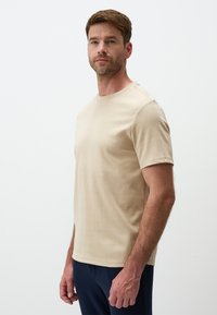 T-shirt beige a girocollo in cotone, maniche corte, vestibilità slim, texture liscia, esposta su un modello maschile su sfondo semplice.