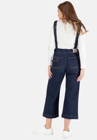 Jeanslatzhosen mit weitem Bein, dunkelblauer Farbe, verstellbaren Trägern und Gesäßtaschen mit genähten Details. Getragen mit weißen Schuhen.