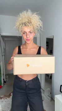 Zalando-mærket papkasse holdt af en person, lys brun med et logo, i et lyst rum med et spejl og tøj i baggrunden.