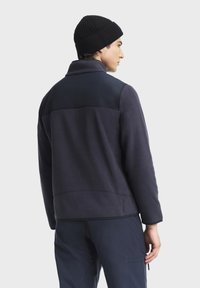 Marinblå fleecejacka med en slät, svart axelpanel, platt krage och subtil branding vid fållen. Bärs med en svart stickad mössa.