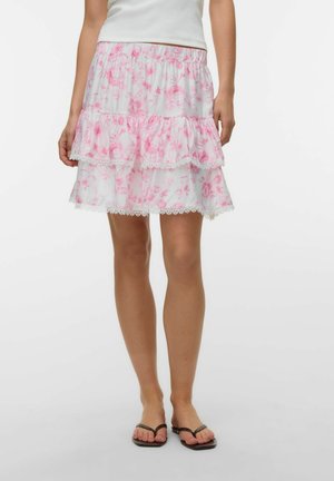 Femme portant une jupe blanche imprimée de fleurs roses avec bordure en dentelle, associée à des sandales noires et un haut blanc, debout sur un fond uni.