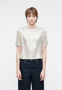 Top recortado de mangas cortas en brillantes lentejuelas plateadas, combinado con pantalones oscuros de mezclilla de tiro alto. Fondo liso que centra la atención en el atuendo.