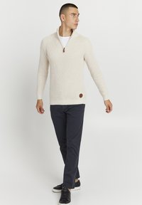 Kremfarget strikkegenser med kvart-zip krage, ribbede mansjetter og hem, sammen med marineblå bukser og svarte joggesko. Brun logo-patch på siden.