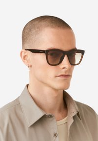 Brown rectangular sunglasses with gradient lenses, thick frames, and subtle logo detail, worn with a light beige shirt, against a white background.