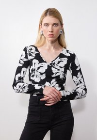 Musthave musta pikema varrukaga topp valge lillemustriga, V-kaelusega disain, soonikuga tekstuur, koos mustade teksadega. Modell poseerib risti kätega.