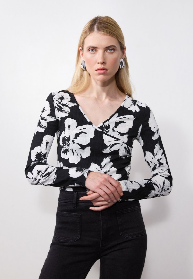 Musthave musta pikema varrukaga topp valge lillemustriga, V-kaelusega disain, soonikuga tekstuur, koos mustade teksadega. Modell poseerib risti kätega.
