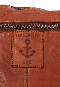 Primo piano di una borsa in pelle marrone con chiusura a zip, caratterizzata da un'etichetta cucita con la scritta "HARBOUR" e un grafico di un'ancora.