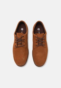 Chaussures en daim brun à lacets avec un bout arrondi et des lacets brun. La semelle intérieure arbore un logo sur un fond brun foncé. Design de semelle plate.