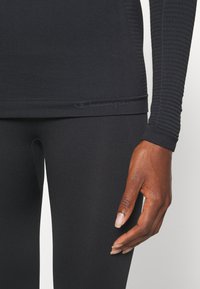 Μαύρο αθλητικό τοπ με μακριά μανίκια, με υφή και εφαρμοστή γραμμή, συνδυασμένο με μαύρες κολάν. Το λογότυπο του Champion είναι εμφανές στο τελείωμα.