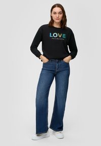 Felpa nera con la scritta "LOVE" in lettere iridescenti e testo sottostante, abbinata a jeans blu a vita alta e gamba larga e sneakers bianche.