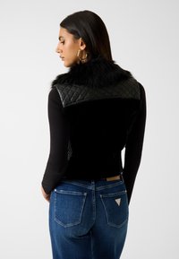 Kobieta w czarnej pikowanej kamizelce z kołnierzem z futerka syntetycznego, czarnym topie z długim rękawem i niebieskich dżinsach, stojąca na tle jednolitego jasnego tła.