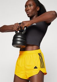 Μαύρη κοντή αθλητική μπλούζα και φωτεινές κίτρινες βερμούδες με μαύρες ρίγες, κρατώντας μια μαύρη kettlebell, μπροστά από έναν απλό φόντο.