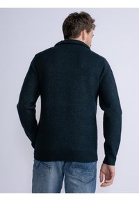 Maglione lavorato a maglia di un blu navy scuro con finitura testurizzata, dotato di colletto a scialle e polsini a costine. Indossato sopra jeans azzurri chiari.