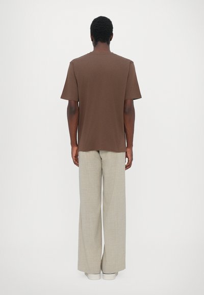 Man with dark skin wearing a brown short-sleeve shirt and light beige wide-leg trousers standing facing a white background.