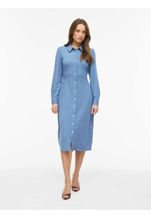 Kvinde iført en lyseblå, knaplukket, knælængde kjole med lange ærmer og krave, samt sorte åbne høje hæle, stående mod en ensfarvet hvid baggrund.