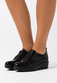 Černé šněrovací boty s texturovaným, lesklým hadím vzorem. Silná černá podrážka s žebrovanými detaily. Kulatá špička a nízký design.