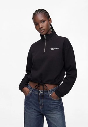 Ung kvinde med flettet hår iført sort lynlåssweatshirt med logo og blå jeans, poserende med hænderne i lommerne mod ensfarvet baggrund.