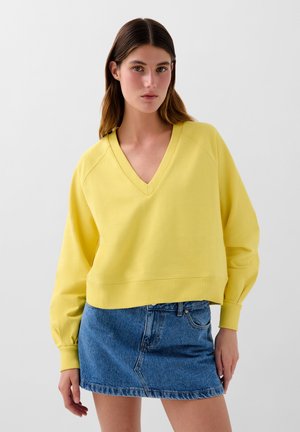 Sudadera amarilla de corte relajado con cuello en V, puños y dobladillo acanalados, combinada con una minifalda de mezclilla. Textura suave, estilo casual.