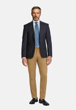 Uomo che indossa un blazer blu navy a righe sottili, camicia azzurro chiaro, cravatta blu con motivo, pantaloni color sabbia e scarpe eleganti nere, in piedi davanti a uno sfondo bianco.