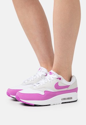 Baskets Nike Air Max blanches et roses portées sur des jambes nues, avec des lacets blancs et des semelles à coussin d'air visibles, sur un fond uni.