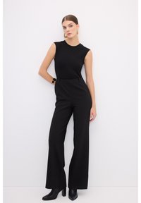 Haut noir sans manches associé à un pantalon noir à taille haute et jambes larges. Tissu lisse ; modèle portant des bottines noires à talons et des bijoux en argent.