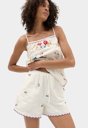 Vrouw draagt een wit geborduurd mouwloos topje en bijpassende korte broek met gekartelde randen en bloemmotieven op een effen achtergrond.