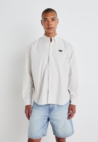 Camicia bianca a righe con un piccolo logo nero, abbinata a pantaloni corti in denim blu chiaro. La camicia ha maniche lunghe e colletto classico.