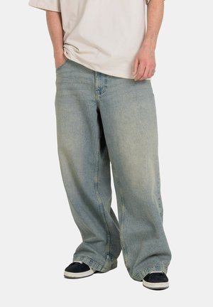 Person wearing loose, light-wash baggy jeans with hands in pockets, standing in black and white sneakers against a plain background.