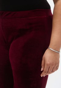 Burgundi bársony leggings sima textúrával és szoros illeszkedéssel, lapos derékpánttal és testhezálló sziluettel. Egy ezüst karkötő is látható.