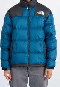 Person iført en blå og sort polstret North Face jakke med frontlynlås og to sidelommer, parret med grå bukser.