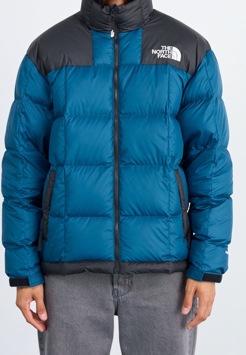 Person iført en blå og sort polstret North Face jakke med frontlynlås og to sidelommer, parret med grå bukser.