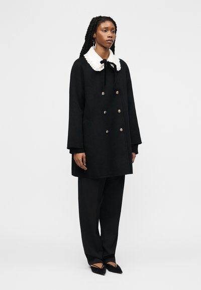 Γυναίκα που στέκεται φορώντας μαύρο παλτό με διπλό πέτο, με λευκό κομψό γιακά και μαύρο παντελόνι φαρδύ, σε απλό φόντο.