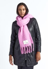 Vrouw die een zwarte leren jas en een oversized zachte roze sjaal met franjes en een "maze" label draagt, staande tegen een witte achtergrond.