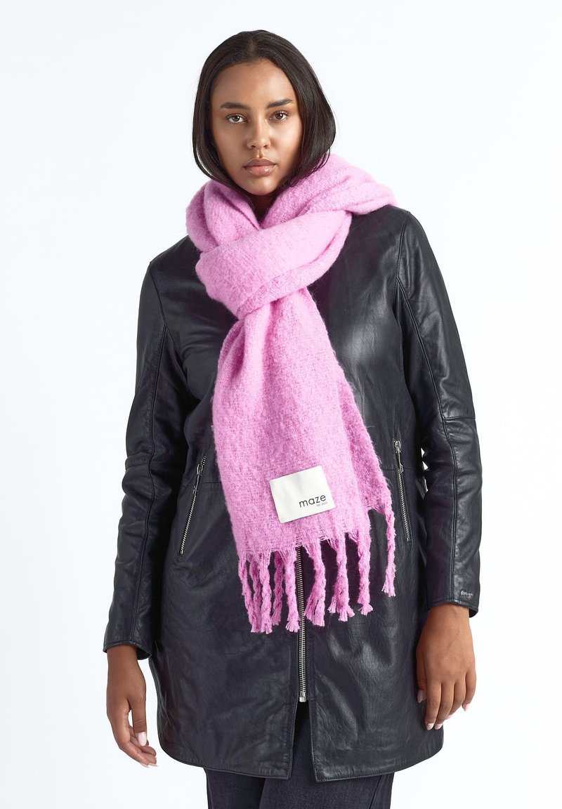 Vrouw die een zwarte leren jas en een oversized zachte roze sjaal met franjes en een "maze" label draagt, staande tegen een witte achtergrond.