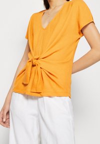 Top naranja texturizado con mangas cortas y escote en V. Detalle de nudo en el frente que añade forma. Usado con pantalones blancos de corte holgado.
