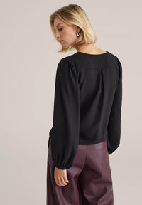 Top nero a maniche lunghe in un tessuto testurizzato, con scollatura tonda e maniche arricciate, abbinato a pantaloni bordeaux a vita alta.