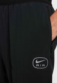 Pantalons de sport noirs en tissu léger avec une taille élastique, dotés d'une poche zippée et d'un logo Nike Air argenté sur la cuisse.