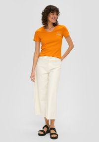 T-shirt en coton orange avec un col rond et des manches courtes, associé à un pantalon blanc à jambe large avec des bords effilochés et des sandales noires.