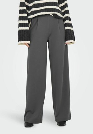 Graue Hosen mit weitem Bein und Falten, aus glattem Stoff, kombiniert mit einem schwarz-weiß gestreiften oversized Pullover. Schwarze Schuhe vervollständigen den Look.