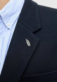 Pin de solapa de pluma plateada sobre un blazer de terciopelo azul marino, combinado con una camisa de rayas azul claro debajo.