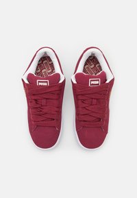 Bordo suede sportbačiai su baltais padais ir raišteliais, turintys raštuotą vidpadį ir logo žymę ant liežuvėlio.