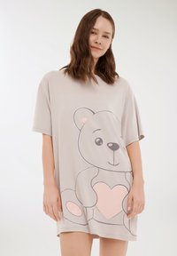 Giovane donna con capelli castani ondulati che indossa un abito beige oversize con un grande orsetto di peluche che tiene in mano un cuore rosa.